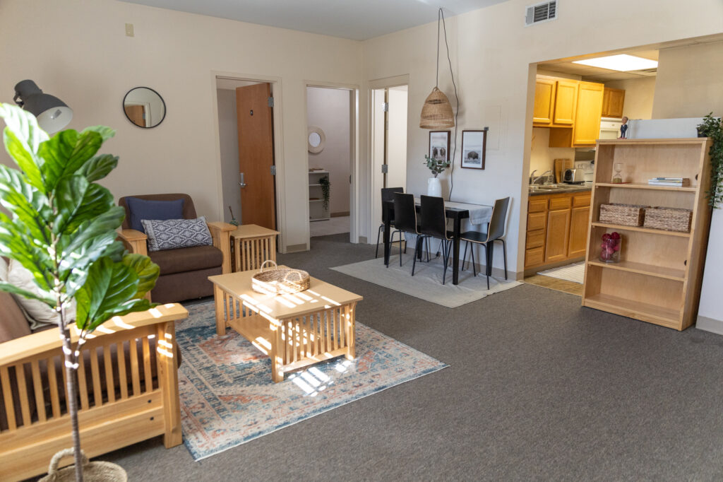 A living room with wooden furniture, a plant, a small dining area, and a view into a kitchen. There are beige walls, carpeted floors, rugs, and doors leading to other rooms. Natural light brightens the space.
