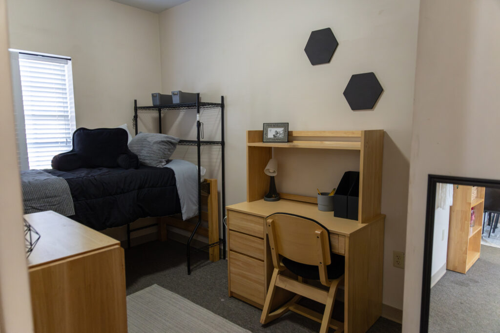 A small dorm room with a single bed, metal shelf, wooden desk and chair, lamp, framed photo, hexagonal wall decor, and a mirror reflecting part of the room. Window blinds let in natural light.