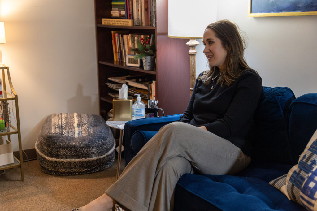 A woman sits on a blue couch in a well-lit room with bookshelves, a lamp, and a patterned ottoman. She is facing to the right, appearing engaged in conversation.