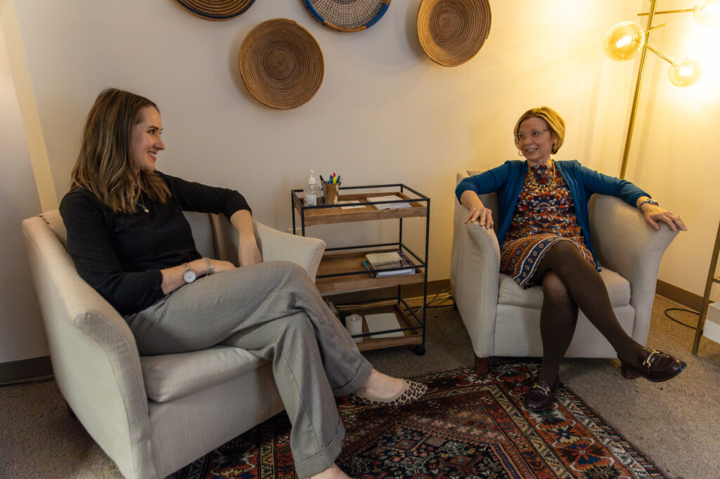 Two women sit in armchairs facing each other in a well-lit room decorated with woven wall art. A small cart with papers, a notebook, and a lamp are visible between them. Both appear engaged in conversation.