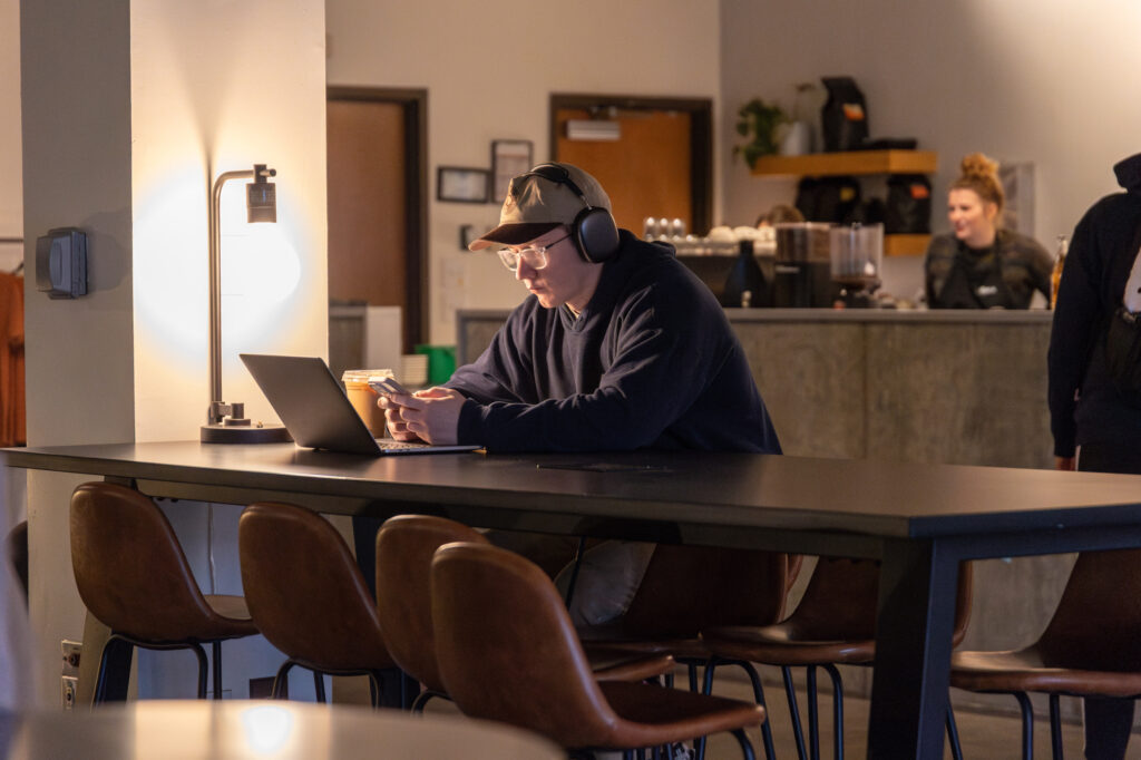 A person wearing headphones and a cap sits alone at a long table in a cafe, looking at a laptop and holding a drink. The background shows a counter, coffee machines, and two people near the barista area.