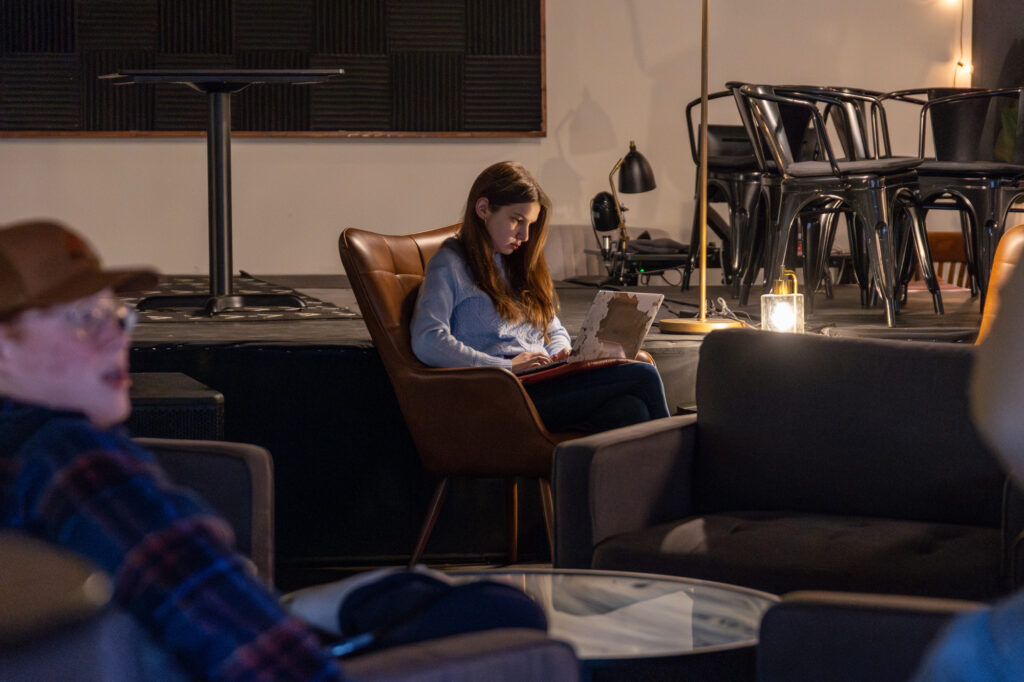 A woman sits in a brown armchair reading a book in a dimly lit room with empty chairs and tables. Other people are seated in the foreground, with a man in a cap partially visible on the left side of the image.