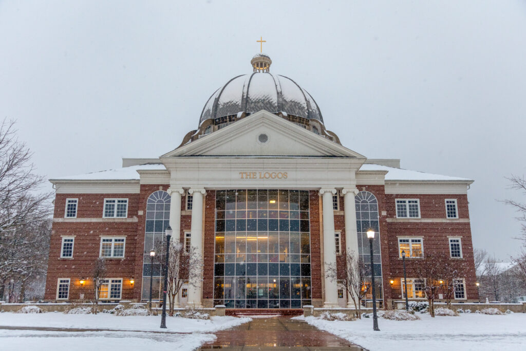 A large brick and glass building with a domed roof and a cross on top, labeled The Logos, stands in snowy weather, with bare trees and lampposts along a paved path leading to the entrance.