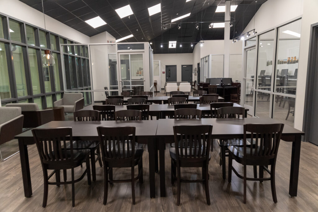 A modern Writing Center room with three dark wooden tables aligned in the center, surrounded by chairs. Glass walls border the space, offering views of additional offices and seating areas. Ceiling lights illuminate the room.