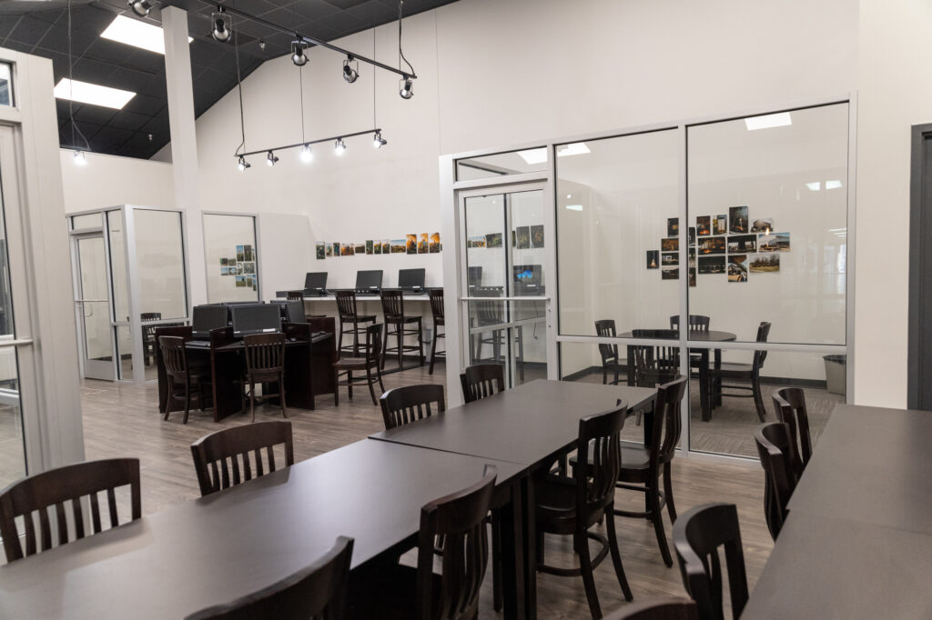 A modern workspace with several dark wooden tables and chairs, computer stations against glass walls, and photos displayed on the walls inside private rooms. The space is well-lit with overhead track lighting, making it an ideal setting for a Writing Center that fosters creativity and productivity.