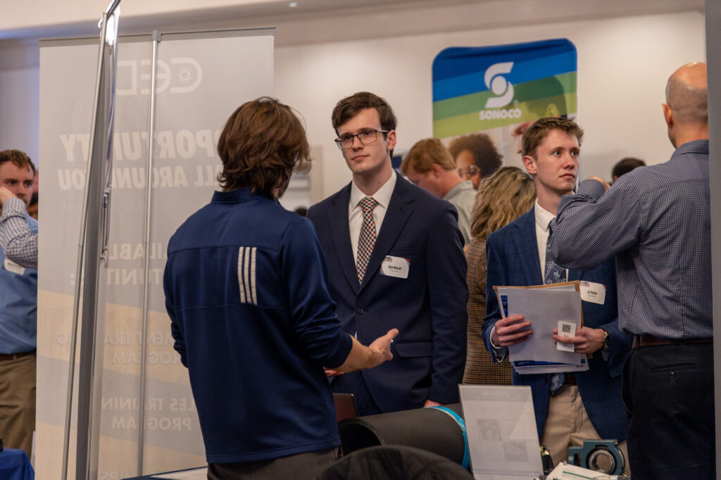 Several people in business attire converse at a career fair or networking event organized by the Vocatio Center. Name tags are visible on some individuals, and informational booths and displays focused on Life Calling and career opportunities can be seen in the background.