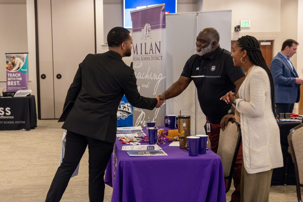 A man in a suit shakes hands with a man behind a table covered in a purple tablecloth at a job fair hosted by the Career Center, while a woman stands nearby smiling. Informational materials and decorations are on the table.