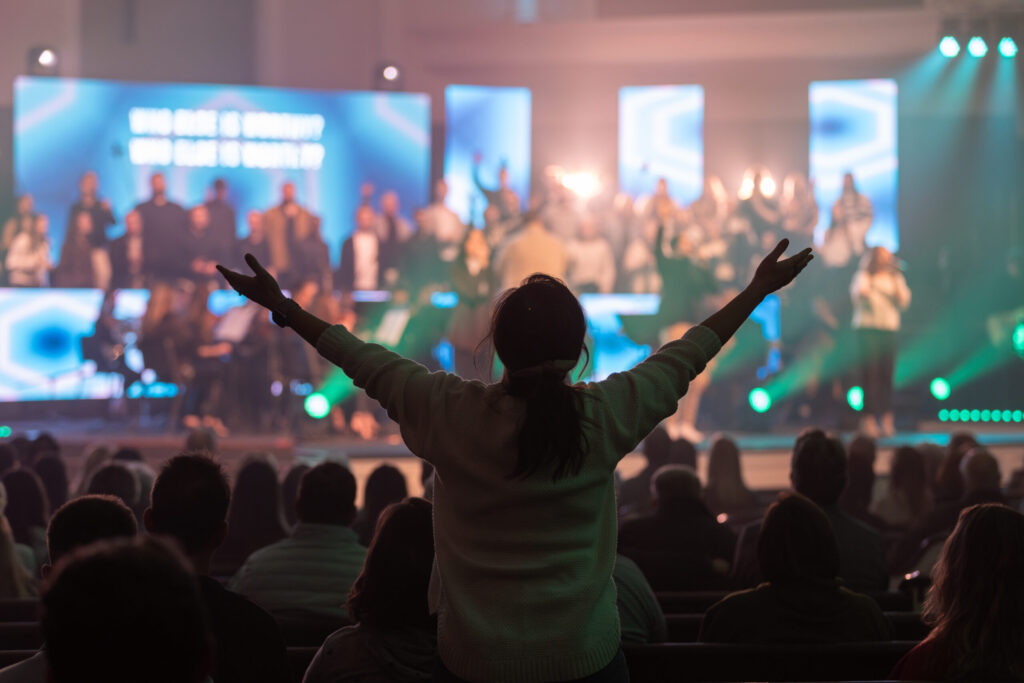 A person stands with arms raised in a crowded auditorium facing a brightly lit stage, where a choir or group of people is performing in front of large screens and stage lights. Audience members are seated.