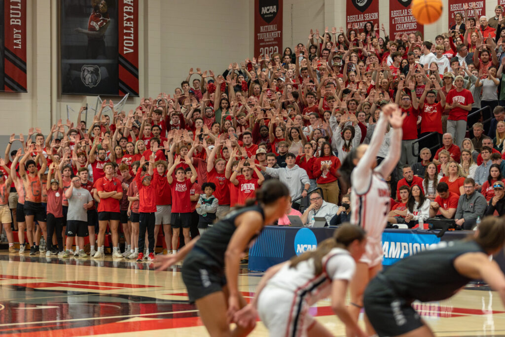 A large crowd of fans, many wearing red, cheer with their arms raised at an indoor basketball game as players prepare for a free throw. Banners and team logos are visible in the background.