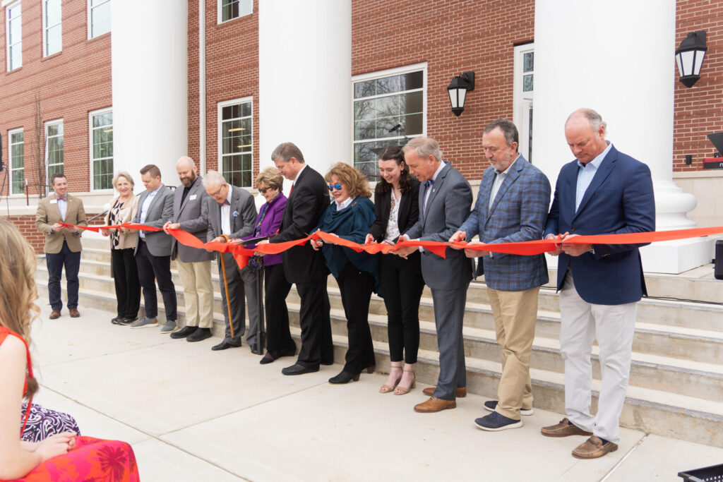A group of people standing in a line cut a large red ribbon with scissors in front of a brick building with white columns, marking a ribbon-cutting ceremony.