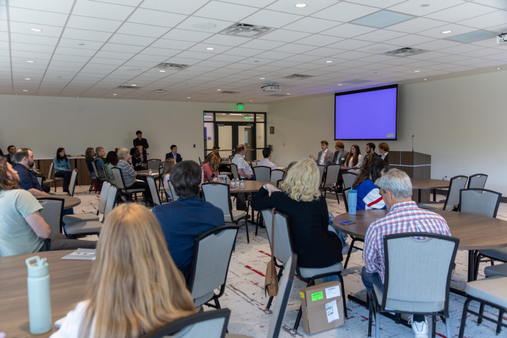 People sit at round tables in a conference room, watching a panel of speakers at the front of the room. The screen behind the panel is turned on but shows a blank blue display.