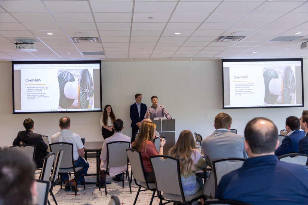 Three people stand at a podium presenting to an audience in a conference room. Two screens display a slide titled Overview with images of airplane turbine parts and bullet points. Attendees are seated, listening to the presentation.