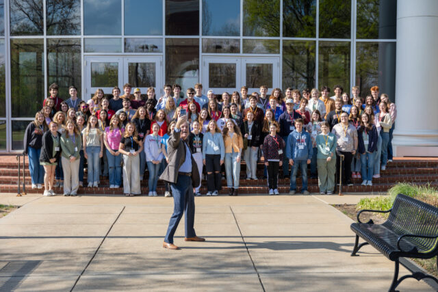 A large group of people is gathered on steps outside a building with glass doors and windows. One person stands in front holding up a phone, appearing to take a selfie with the group on a sunny day.
