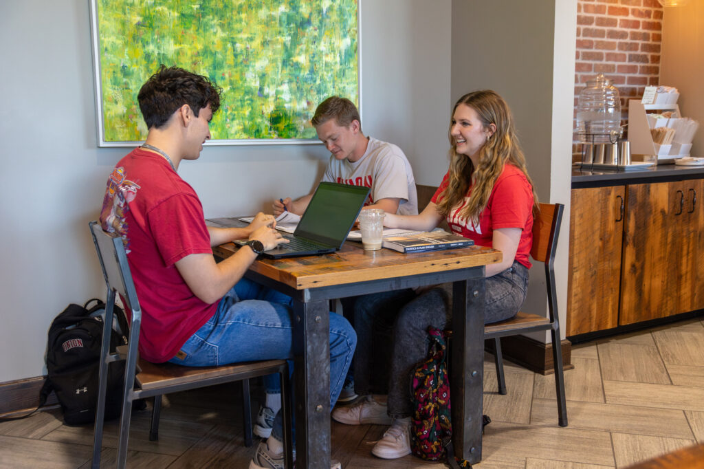Three young adults sit at a wooden table in a café, working on laptops and taking notes. They appear focused and engaged in conversation. A painting and coffee station are visible in the background.