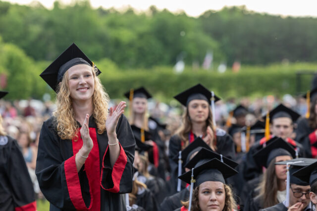 A group of students in black graduation gowns and caps gathers outdoors; one woman in front stands and claps, while others sit and watch the ceremony. Trees and a crowd are visible in the background.