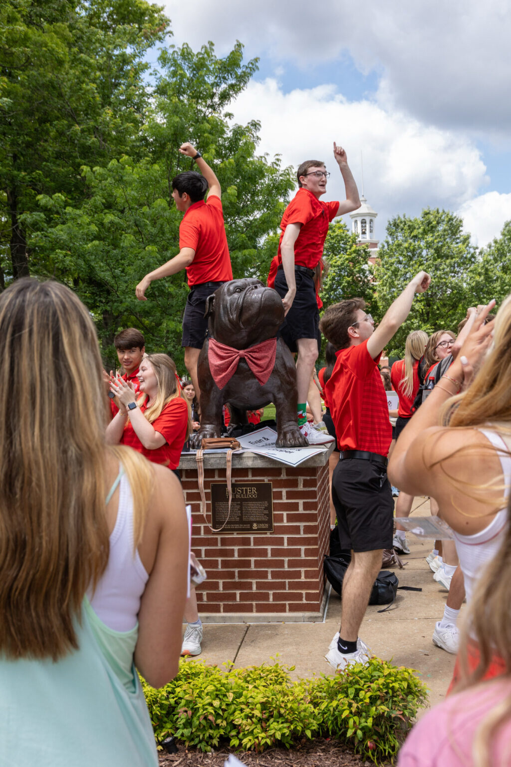 A group of First-Year students in red shirts pose and gesture around a bronze bulldog statue on a brick pedestal outdoors. Some face the crowd, while others stand on the pedestal. Trees and a building are in the background.