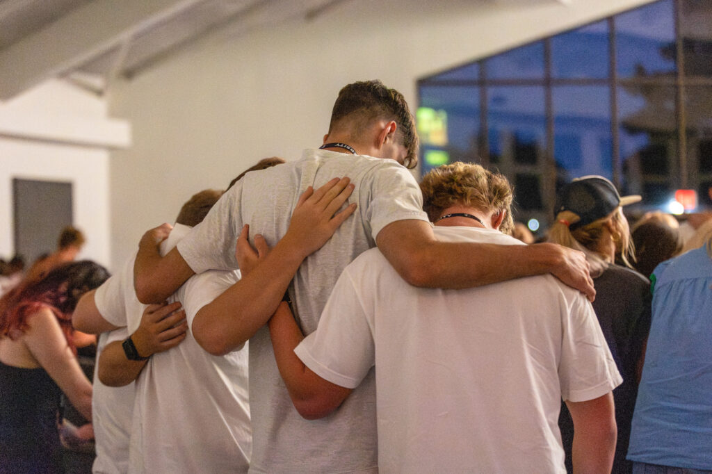 A group of four people, seen from behind, stand closely together with their arms around each other in a well-lit indoor space with large windows.