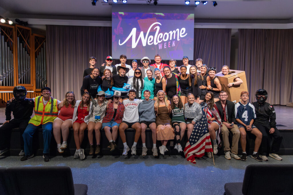 A large group of young adults in costumes poses on a stage in front of a screen that says “Welcome Week.” Some are sitting, some are standing, and a few hold props such as a flag and a cardboard box.