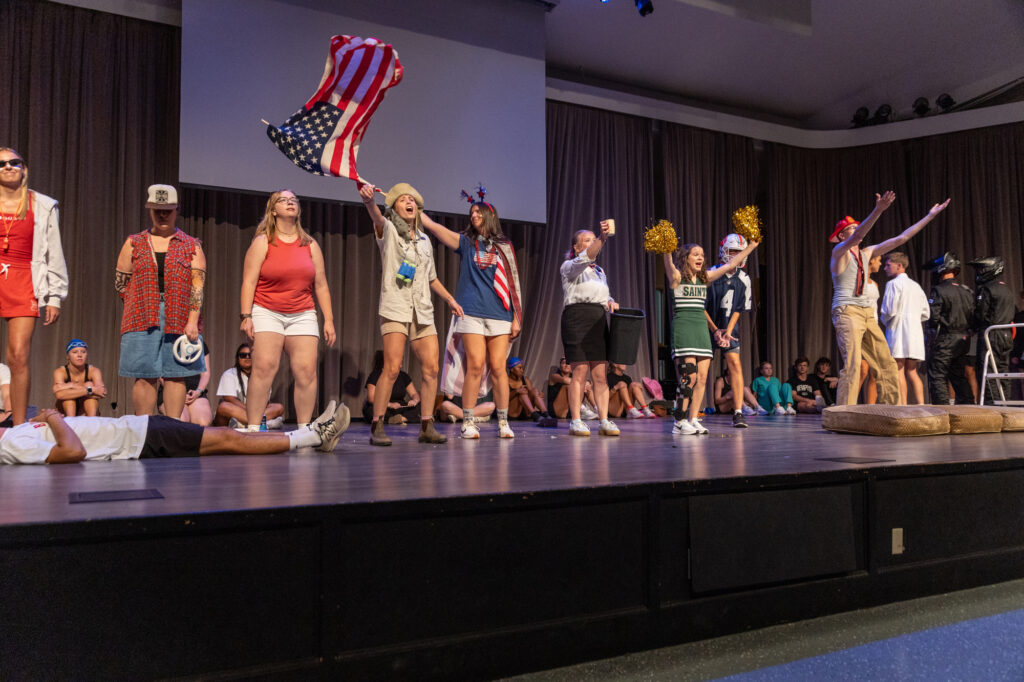 A group of people in various costumes stand and pose on a stage; one person waves an American flag, while others wear sports outfits, hats, and sunglasses. Some people are sitting or lying down, and the background is a curtained wall.
