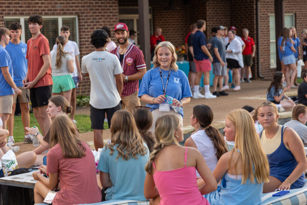 A group of young adults socializes outdoors near a brick building. Some are sitting on benches, while others stand and chat. Most wear casual summer clothing. One person in a blue shirt with a nametag stands near the center.