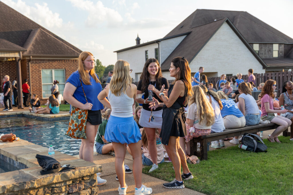 A group of people socializes beside a backyard pool at a gathering. Some stand talking near the pool, while others sit on benches or the grass. Houses and a wooden fence are visible in the background.