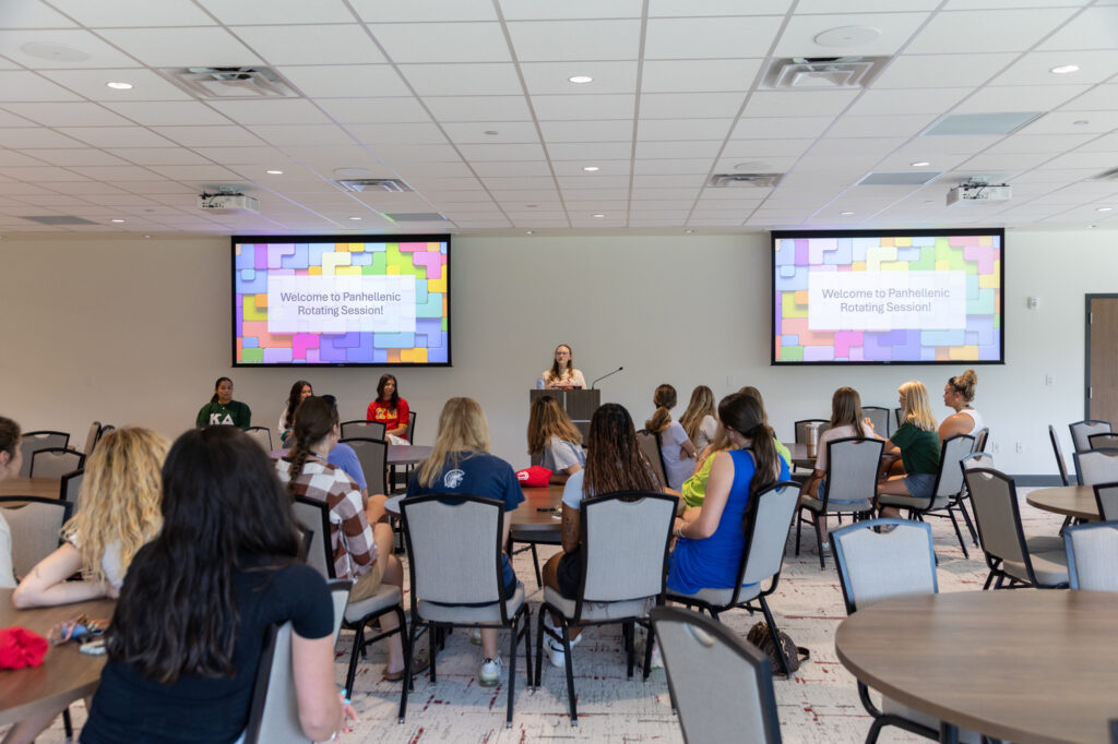 A group of people sit at round tables facing a speaker at a podium in a conference room. Two screens behind the speaker display the message Welcome to Panhellenic Rotating Session!.