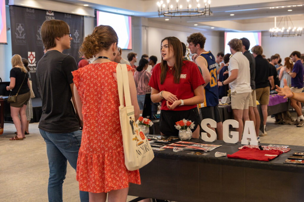 A woman stands behind a table with SGA letters, talking to students at a campus event. There are brochures, flowers, and a shirt on the table. Other people are seen mingling in the background.
