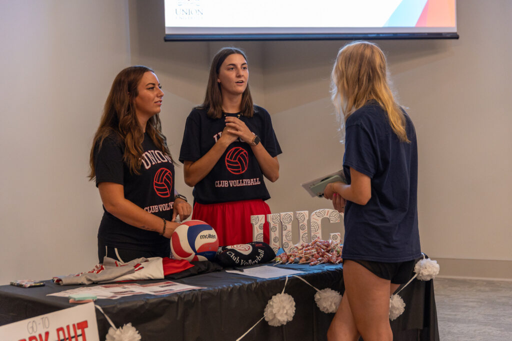 Three people stand at a table with volleyball-themed items and club flyers in a well-lit room. Two are behind the table talking to a third, who is holding papers. The table has pom-pom decorations and a volleyball.