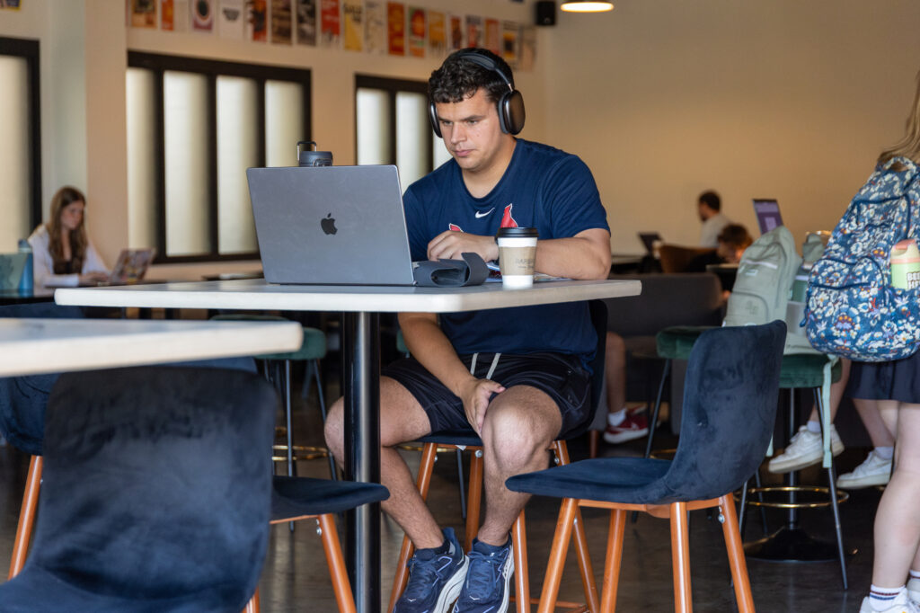 A man wearing headphones sits at a table in a cafe, looking at his laptop. There is a coffee cup and a water bottle on the table. Other people are visible in the background working on laptops.
