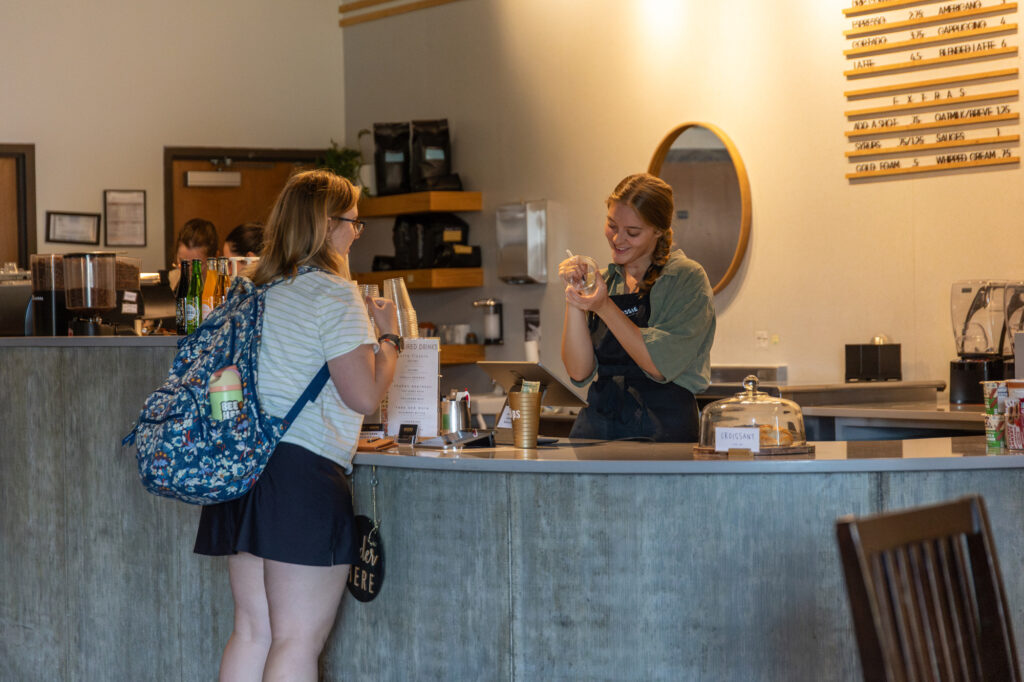 A customer with a backpack talks to a barista behind the counter in a coffee shop. The barista holds a glass, smiling, while various items like cups and a display case are visible on the counter.