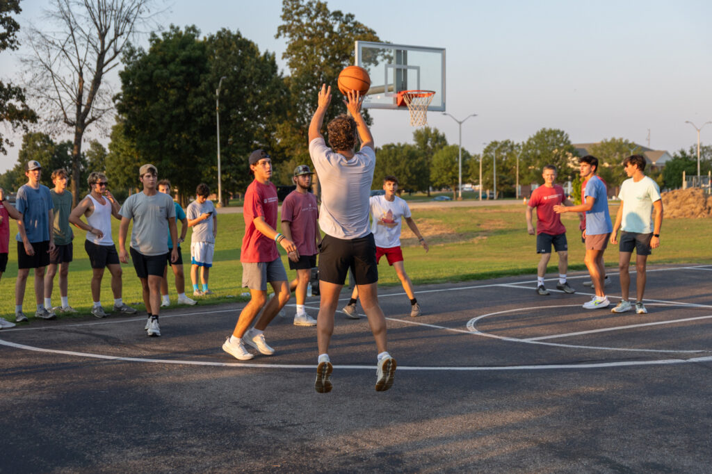 A group of young men, part of the Residence Life community, play basketball on an outdoor court. One player jumps to take a shot while others watch or get ready to rebound. Trees and park scenery are visible in the background.