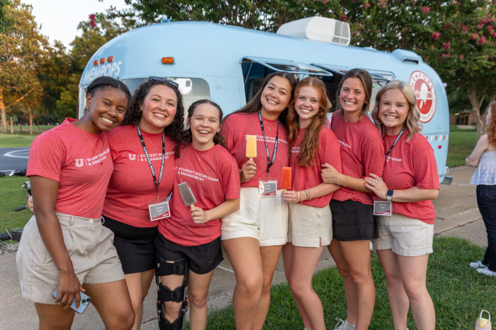 Seven young women in matching red shirts and name badges stand together smiling outside, in front of a blue blood donation trailer and trees, holding what appear to be folded T-shirts or hand towels.