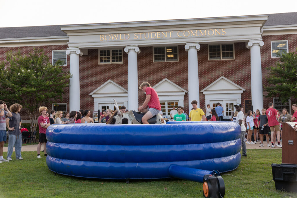 A person rides a mechanical bull in an inflatable ring in front of Bowld Student Commons as a crowd watches outdoors, enjoying one of the many Residence Life events. The brick building has white columns and windows.