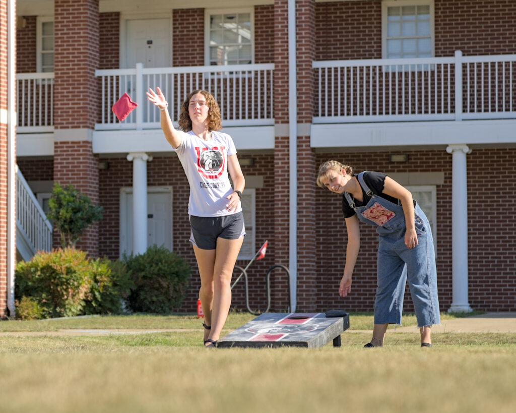 Two people play cornhole outside on a grassy lawn in front of a brick building. One person tosses a red bean bag, while the other watches. The weather appears sunny and clear.