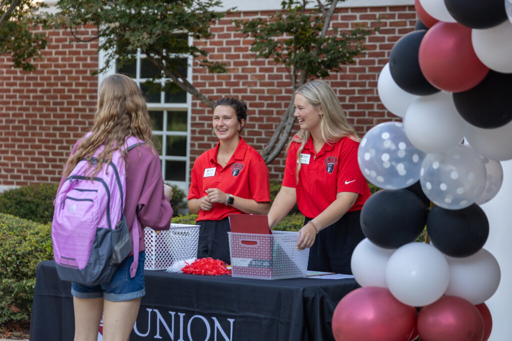 Two women in red shirts stand behind a registration table outdoors, talking to a student with a pink backpack. Black, white, and red balloons decorate the table. A brick building and greenery are in the background.