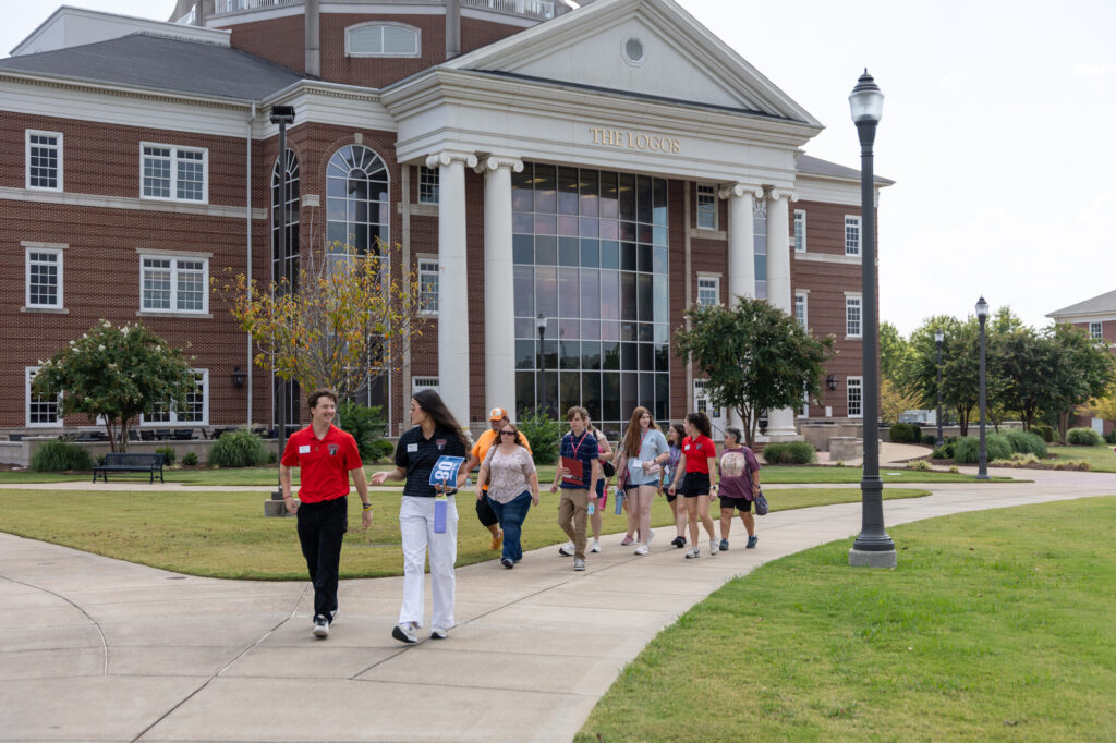 A group of people walk on a sidewalk in front of a large brick building with columns and glass windows. The building has a sign that says The LCOS. The area is landscaped with grass and trees.