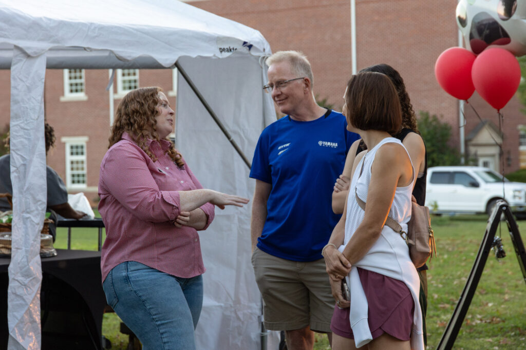 Four adults, including current parents of Union University students, stand talking outside near a white tent, a table with food, and red balloons. One woman gestures while speaking, and the group listens. A brick building and a white vehicle are visible in the background.