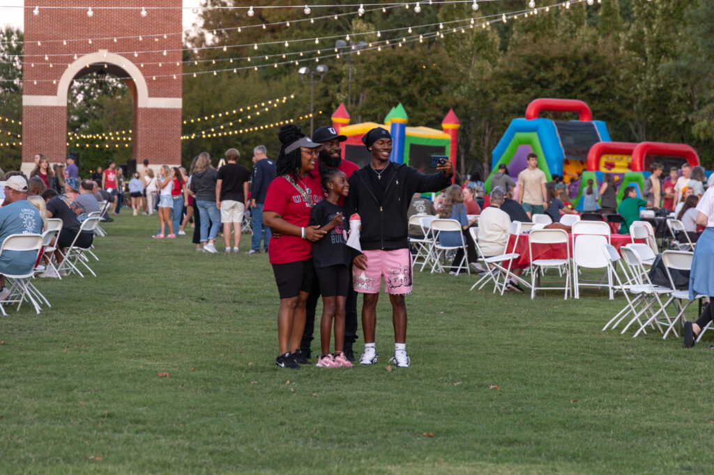 Three people stand together for a photo at an outdoor event with tables, chairs, and inflatable play structures. Other attendees—including current parents of Union University students—are visible in the background, and string lights are hung overhead.