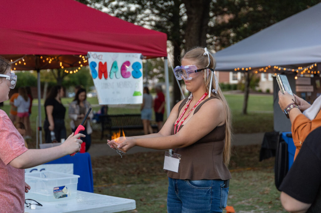 A woman wearing safety goggles holds a small flame in her hand with assistance from another person using a lighter at an outdoor science demonstration event, with tents and a sign in the background.