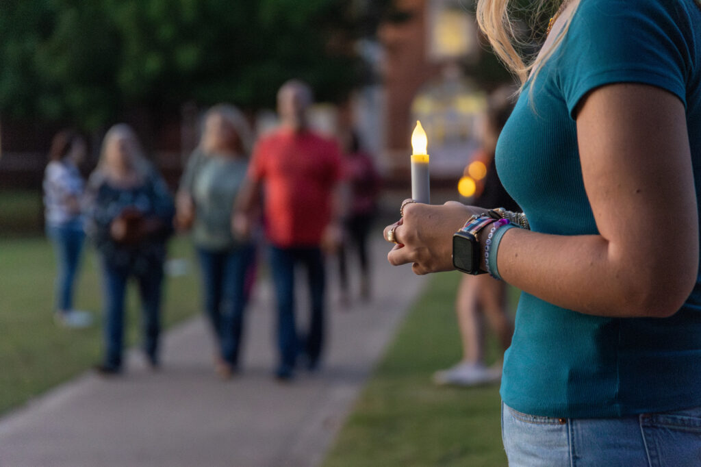 A person holding a lit candle stands in the foreground, symbolizing hope and unity, while a group of people walks on a sidewalk in the background. The scene takes place outdoors in the evening, evoking themes of social justice and just & caring communities coming together.