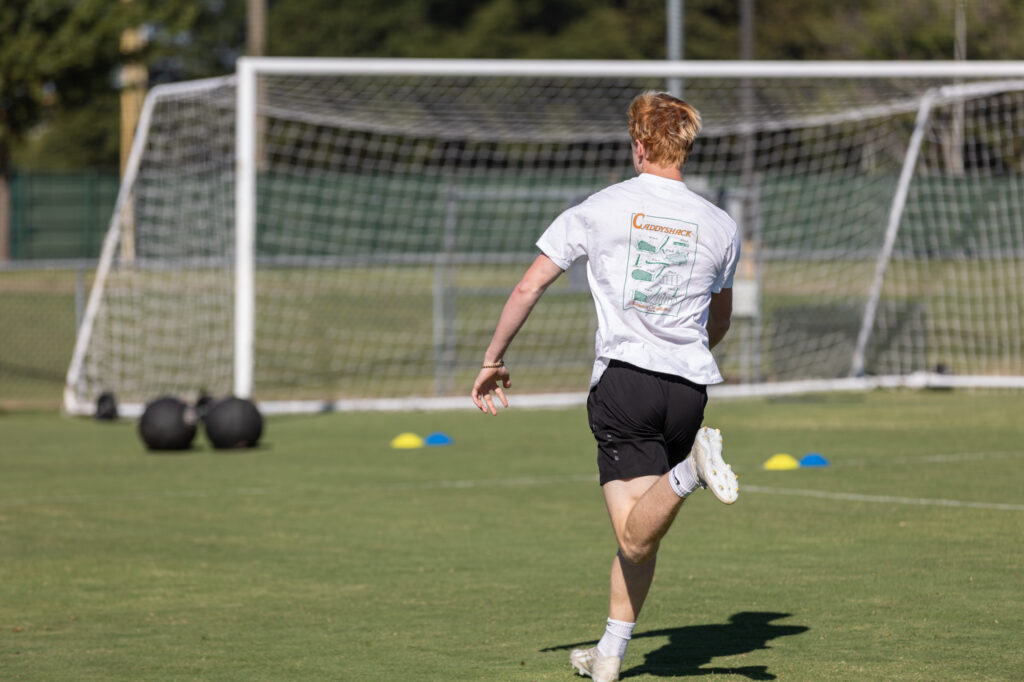A person with light hair wearing a white T-shirt and black shorts runs on a grass field toward a soccer goal, with medicine balls and cone markers visible in the background.