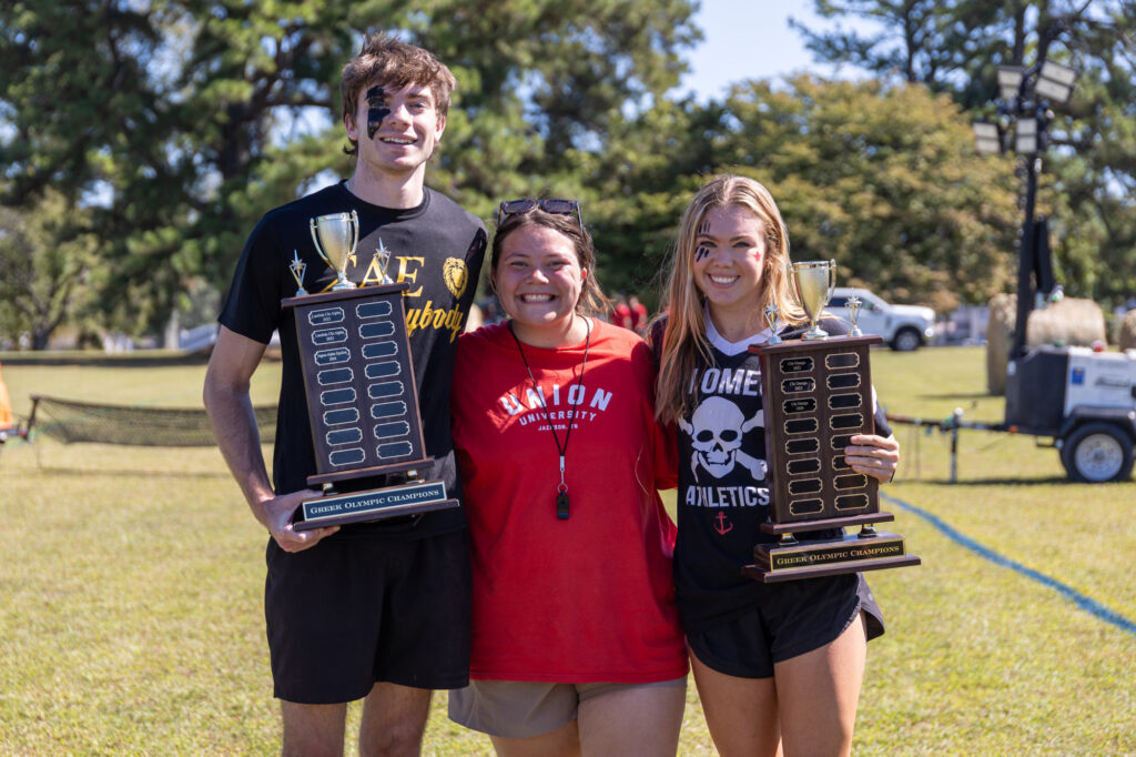 Three people stand on grass outdoors. The two on the sides hold large trophies and wear athletic clothes; the person in the center wears a red shirt and whistle. All three are smiling, with trees and equipment in the background.