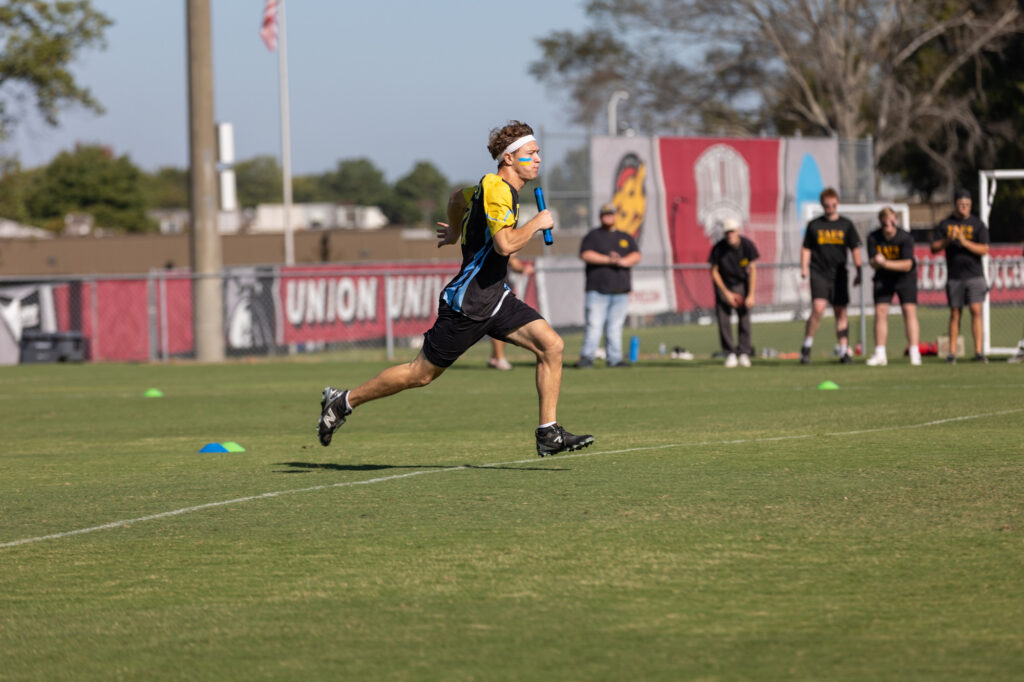 A runner in athletic gear holds a blue baton while sprinting on a grassy field during a relay race. Several people stand in the background near a red fence and sports banners.