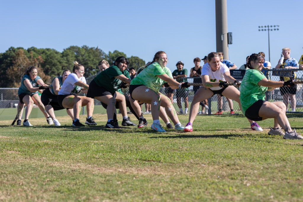A group of people participate in a tug-of-war on a grassy field, pulling on a rope in opposite directions, with trees, a fence, and spectators in the background under a clear sky.