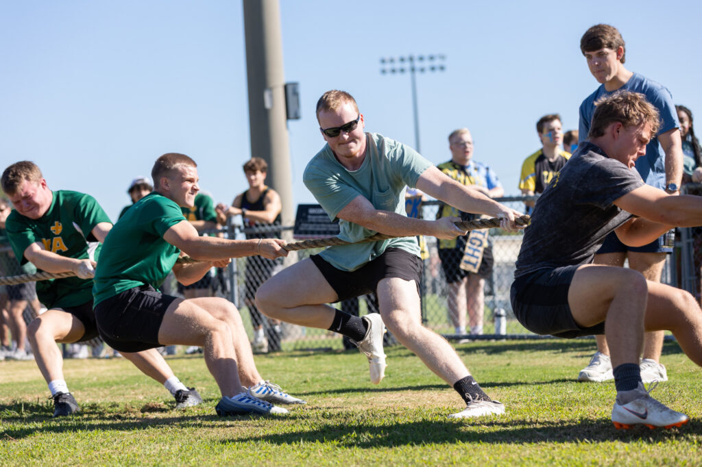 A group of men are playing tug-of-war outdoors on a grassy field. Some are wearing green shirts and others are in dark colors. Spectators watch in the background under a clear, sunny sky.