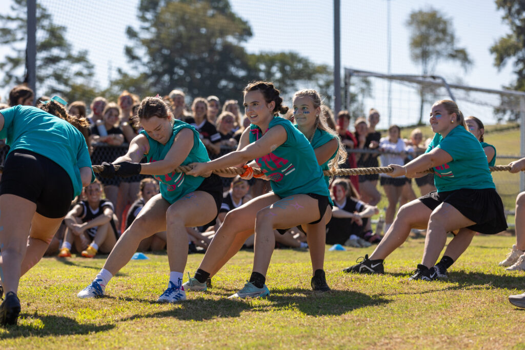 A group of young women in teal shirts participate in a tug-of-war competition outdoors, pulling on a rope with a crowd of onlookers in the background. The event takes place on a sunny day.