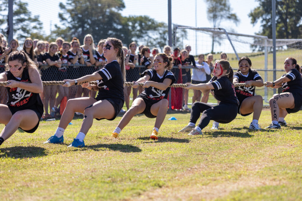 Six young women in matching shirts participate in a tug-of-war outdoors, pulling on a rope. A crowd watches from behind a fence, and trees and sports equipment are visible in the background.