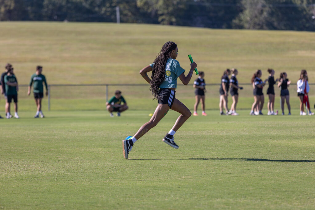 A runner holding a baton sprints across a grassy field during a relay race, with other people standing and watching in the background.