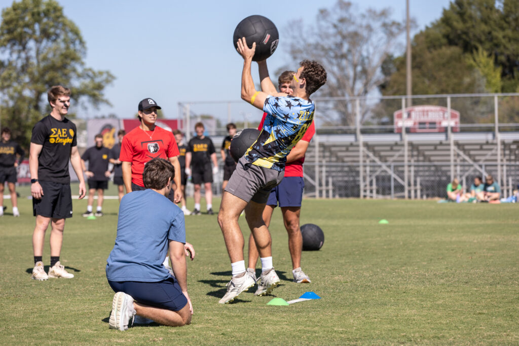 A group of young men are outdoors on a field. One person lifts a large black ball above his head while others watch. Some wear T-shirts and shorts. There are cones and more black balls on the grass. Bleachers are in the background.