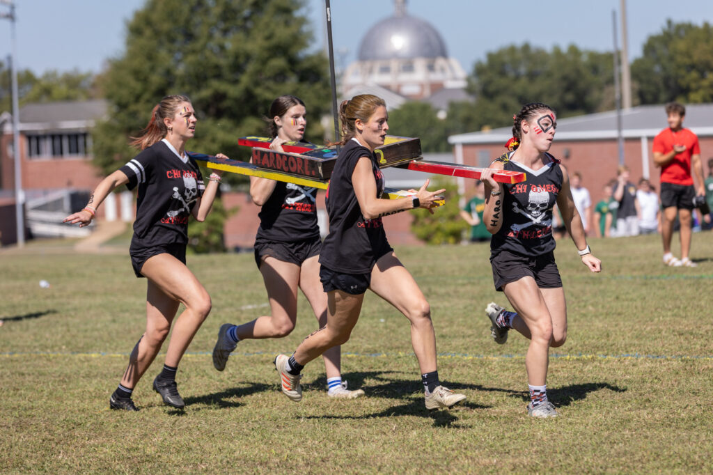 Four women in black shirts and shorts run outdoors on grass, carrying a decorated wooden structure together. One woman has face paint. A brick building and dome are in the background. Spectators are visible in the distance.